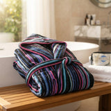 Accappatoio con cappuccio in morbida spugna di cotone a righe colorate fucsia, blu e nere, piegato su una panca in legno in un bagno elegante.
