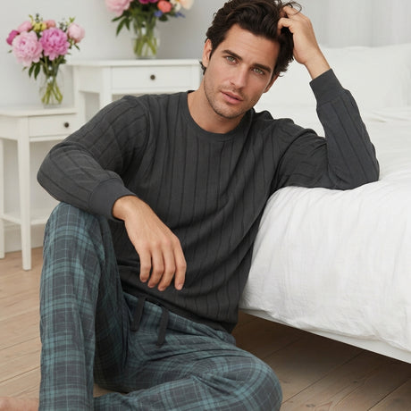 Modello uomo seduto accanto al letto indossa il pigiama invernale in caldo cotone grigio a costine con pantalone a quadri